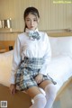 A woman in a white shirt and plaid skirt sitting on a bed.