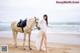 A woman in a white dress standing next to a white horse on a beach.